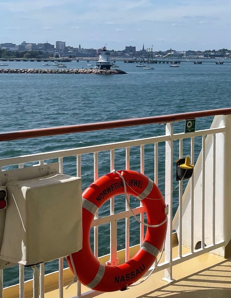 A small lighthouse that resembles a spool of white thread with a wraparound deck near the bottom is in the middle of the frame with a ships railing and Norwegian jewel life ring in the foreground. 