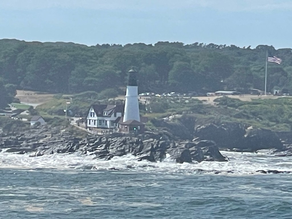 Classic tapered shaped white lighthouse with black tip sits between a small shack like house and a large 3 story classic inn like building as waves pound the rocky shore. 