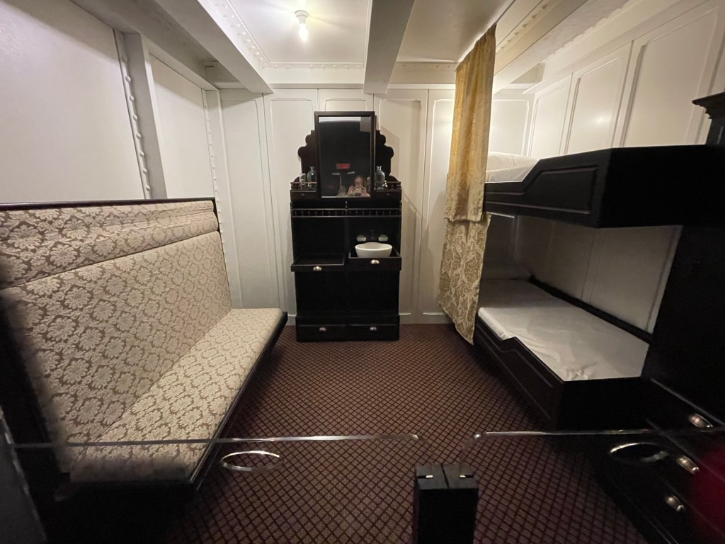 Along left wall is a long and tall wooden bench with upholstered seat and back, along right wall are an upper and lower bunk both dark wood both with soft privacy curtains, plus an armoire, and in the middle a dark wood wash stand with basin. Walks are stark white with white wood trim.