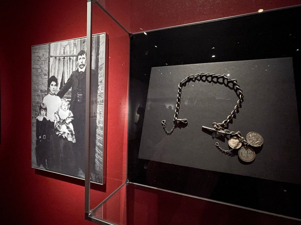 On the wall next to the framed shadow box of a silver chain is a photo of the family, father, mother, and 2 young sons.