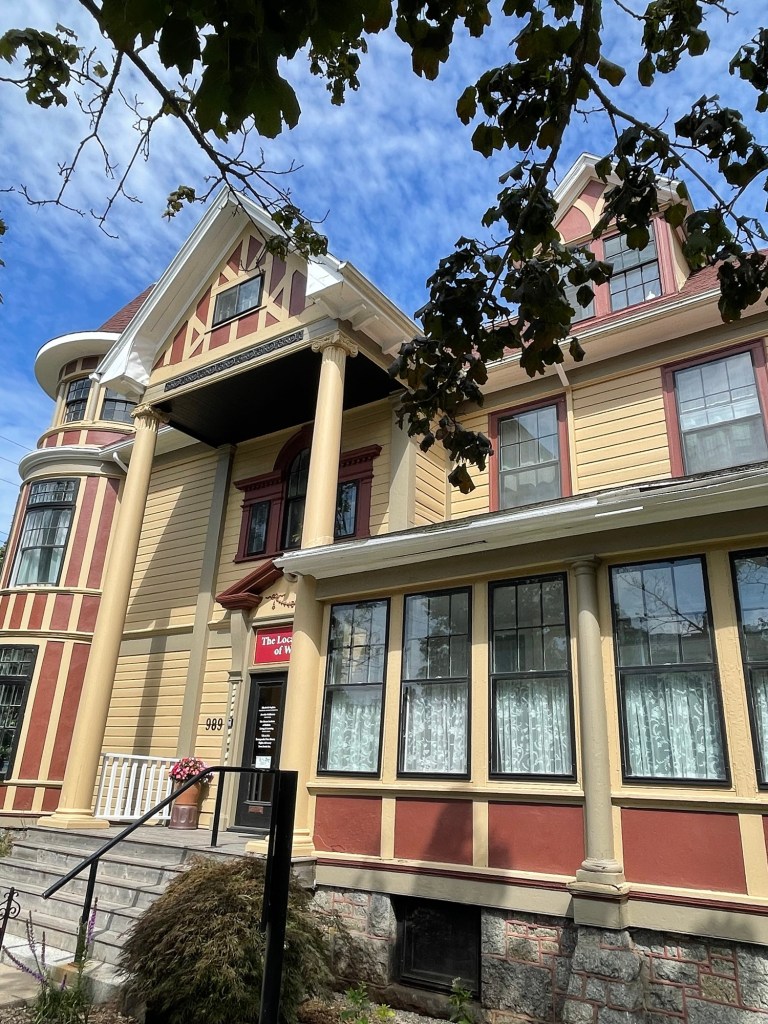 A colorful Queen Anne Revival house with yellow siding, red trim, ornate gables, tall columns, and a rounded turret, partly framed by tree branches against a blue sky.