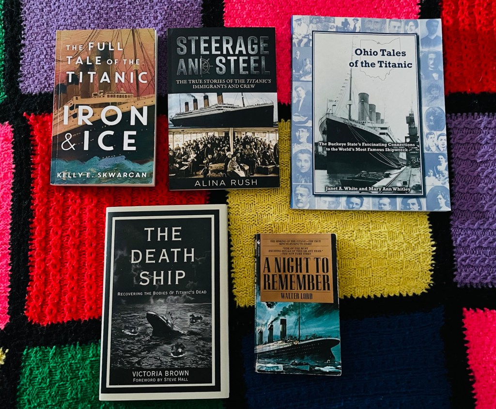 The five books listed in this article are arranged on a thick, hand-knitted blanket made of large, textured squares of bold high contrast colors stitched together in a patchwork pattern.
