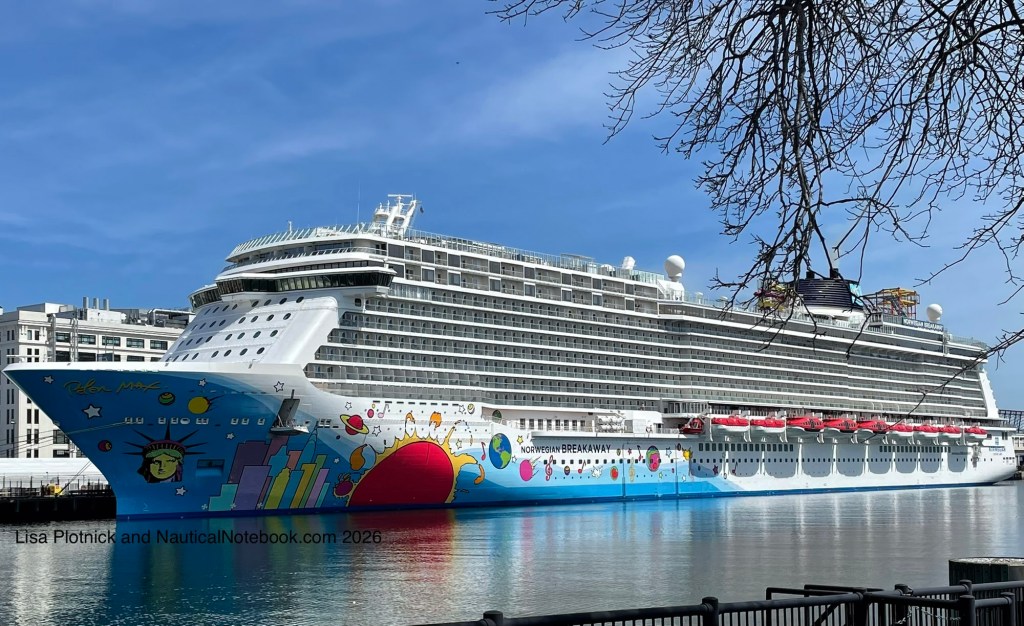 Cruise ship Norwegian Breakaway docked in Boston on a clear day, its hull painted with colorful pop-art graphics including a stylized Statue of Liberty, sun, and skyline. 7 full decks of balconies plus 3 partial forward decks of balconies are above the hull and a multi level winding slide is aft on the top deck. Calm water reflects the ship and bare tree branches frame the scene.