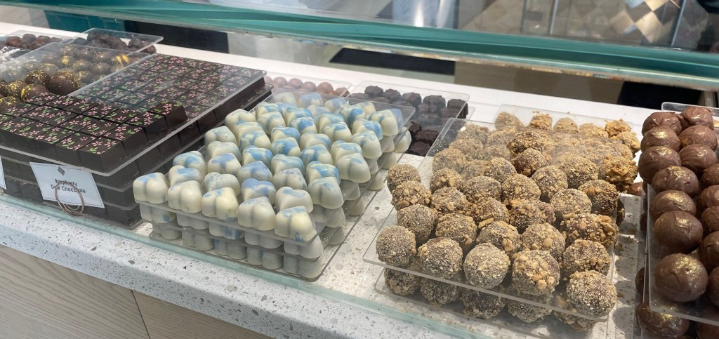  A glass display case filled with assorted handmade chocolates, including white-and-blue molded pieces, chocolate-covered truffles rolled in nuts or crumbs, and round chocolate-covered candies