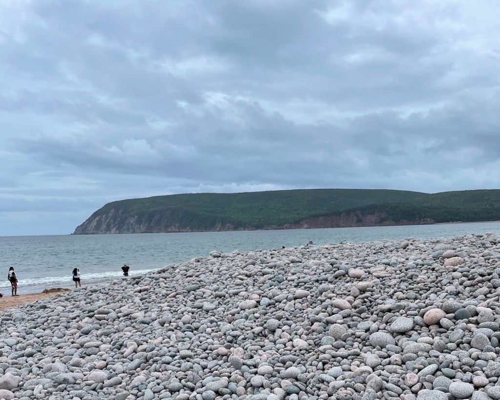 Beyond a beach filled with small rocks, three people in swimwear stand on a strip of sand getting ready to enter a calm ocean as a mild wave comes ashore. 