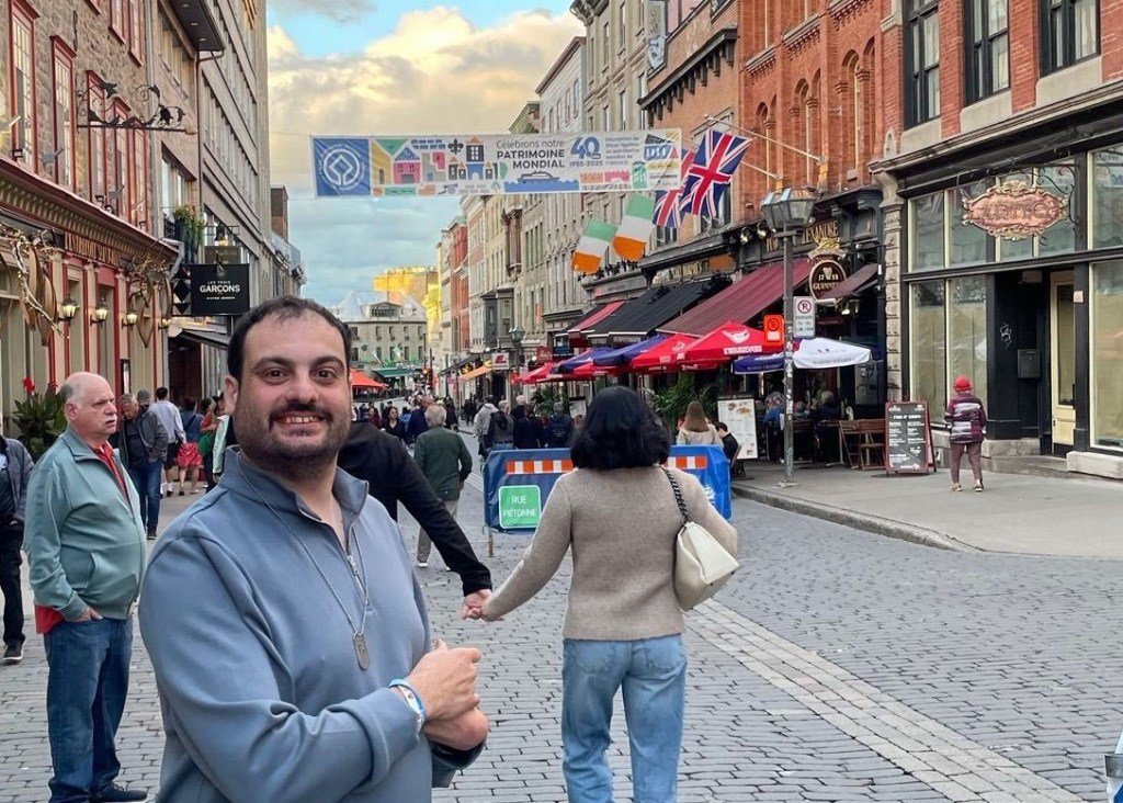 A cobblestone street lined with historic stone and brick buildings, colorful shopfronts, and outdoor restaurant seating. International flags hang above the busy street, and a banner stretches between the buildings. A man in a gray pullover smiles in the foreground beneath a bright blue sky with scattered clouds.
