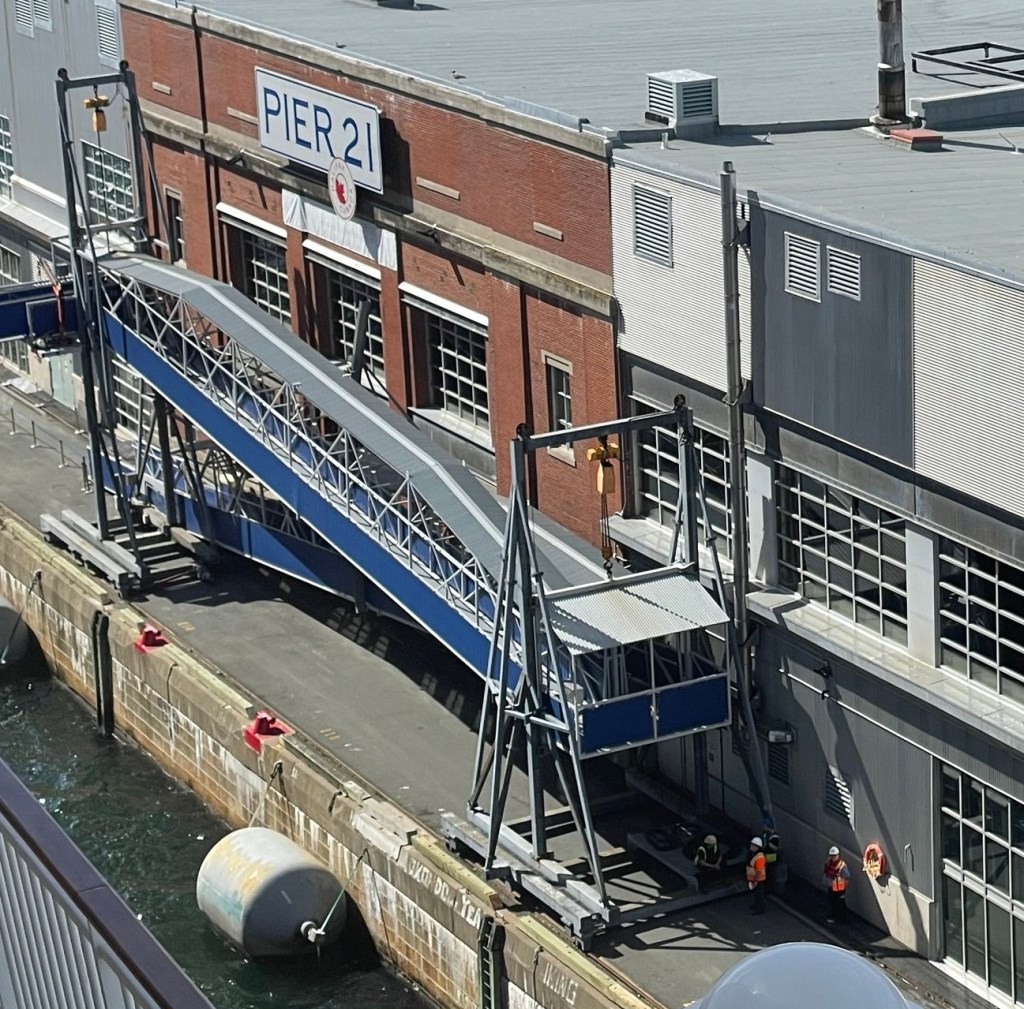  A view from the ship looking down at a brick-and-metal cruise terminal building marked “PIER 21.” A covered blue-and-gray passenger gangway extends from the terminal to the ship.