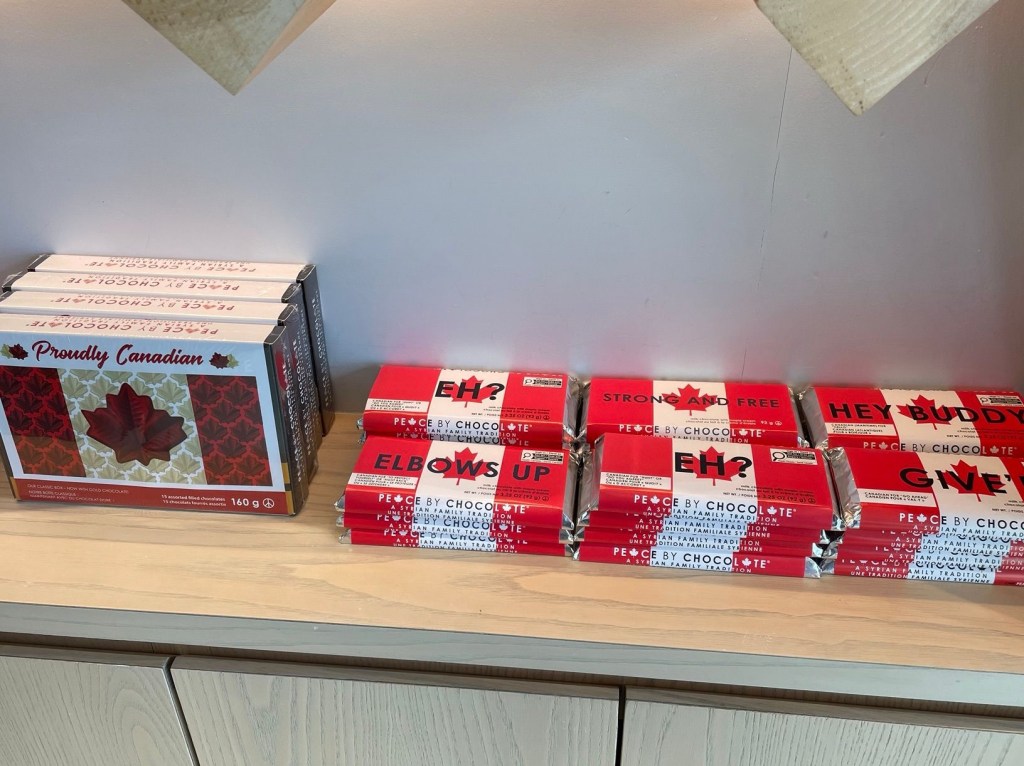 Peace by Chocolate boxed chocolates and large individual bars lined on a shelf. The wrapping on the bars depicts the Canadian flag with labels that include Eh?, elbows up, strong and free, and hey buddy. 