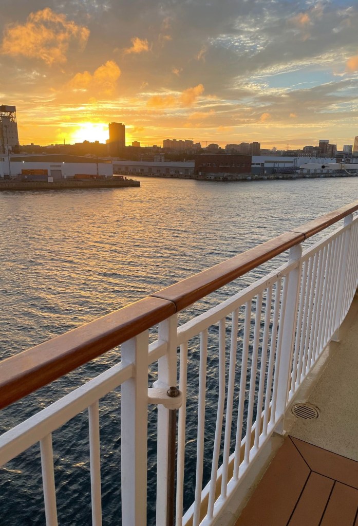 The sun sets over Halifax harbor, casting a golden reflection across the water. The city’s buildings form a dark skyline in the distance, while the ship’s white railing and wooden handrail frame the view in the foreground.