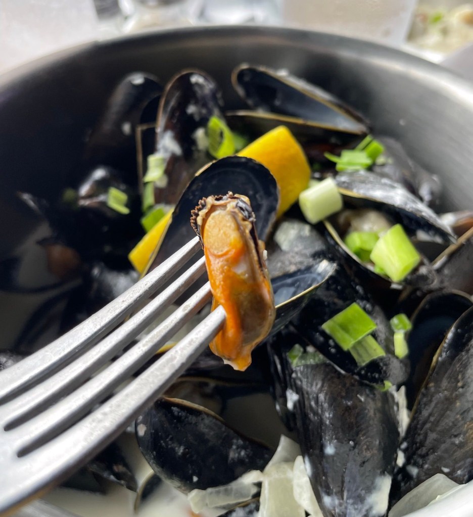 A fork holds a single orange steamed mussel above a black bowl filled with open dark shells, creamy sauce, chopped green onions, and a lemon wedge. Drinking glasses and other dishes are softly blurred in the background.
