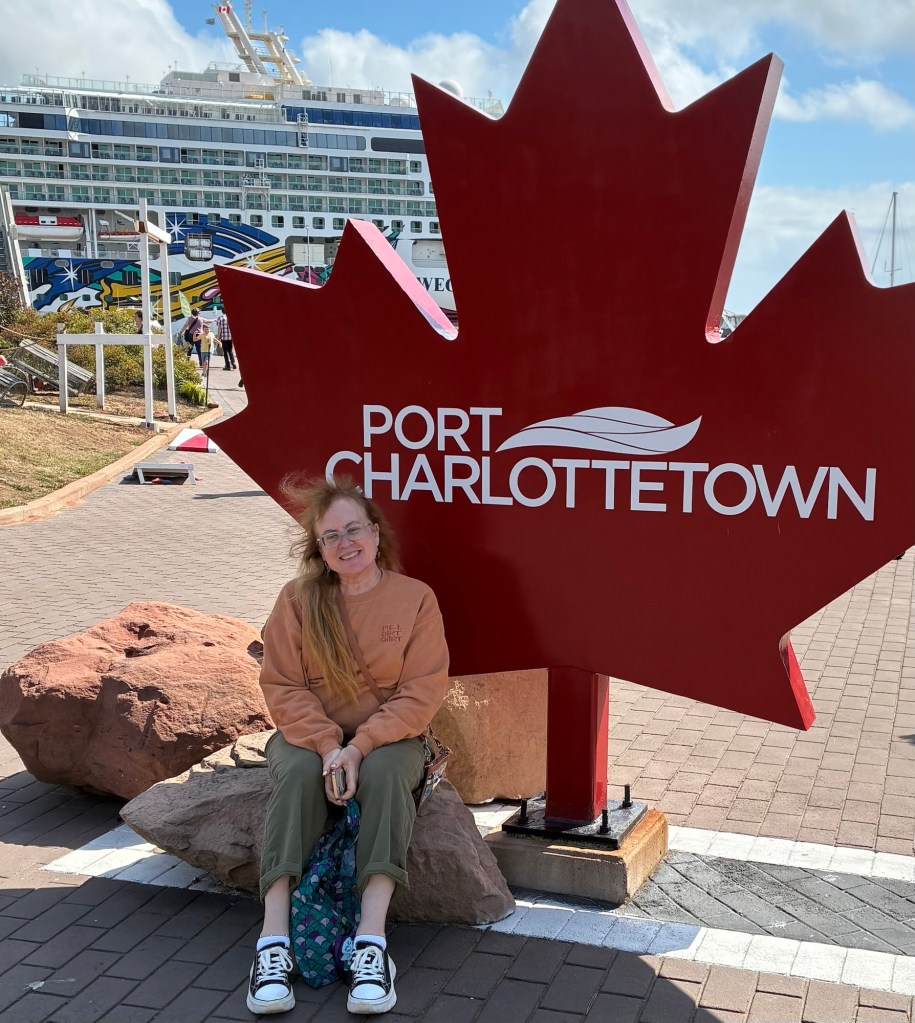 Lisa, a Caucasian woman with long brown hair, sits next to a maple leaf shaped sign with the words Port Charlottetown on it, with a cruise ship in the background. She wears a reddish orange crew neck sweatshirt that’s the same color as the large rocks she sits next to.
