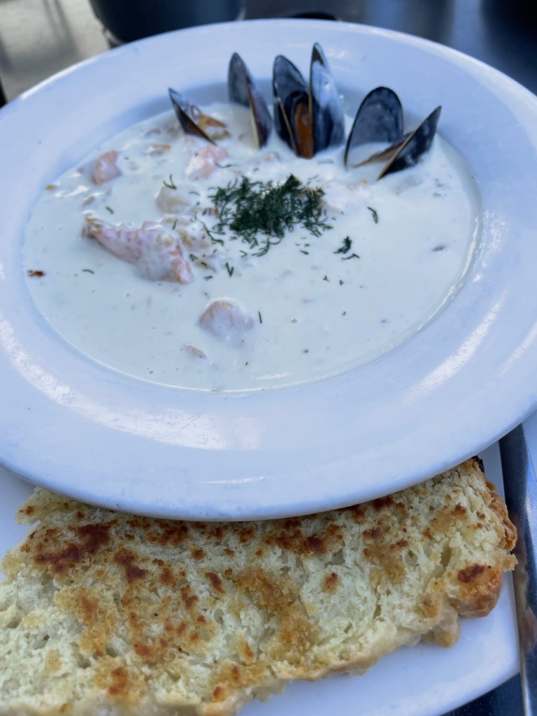A bowl of creamy white seafood chowder topped with fresh dill, with mussels in their shells and pieces of fish, shrimp, and scallops visible in the broth. A large piece of toasted bread sits beneath the bowl.