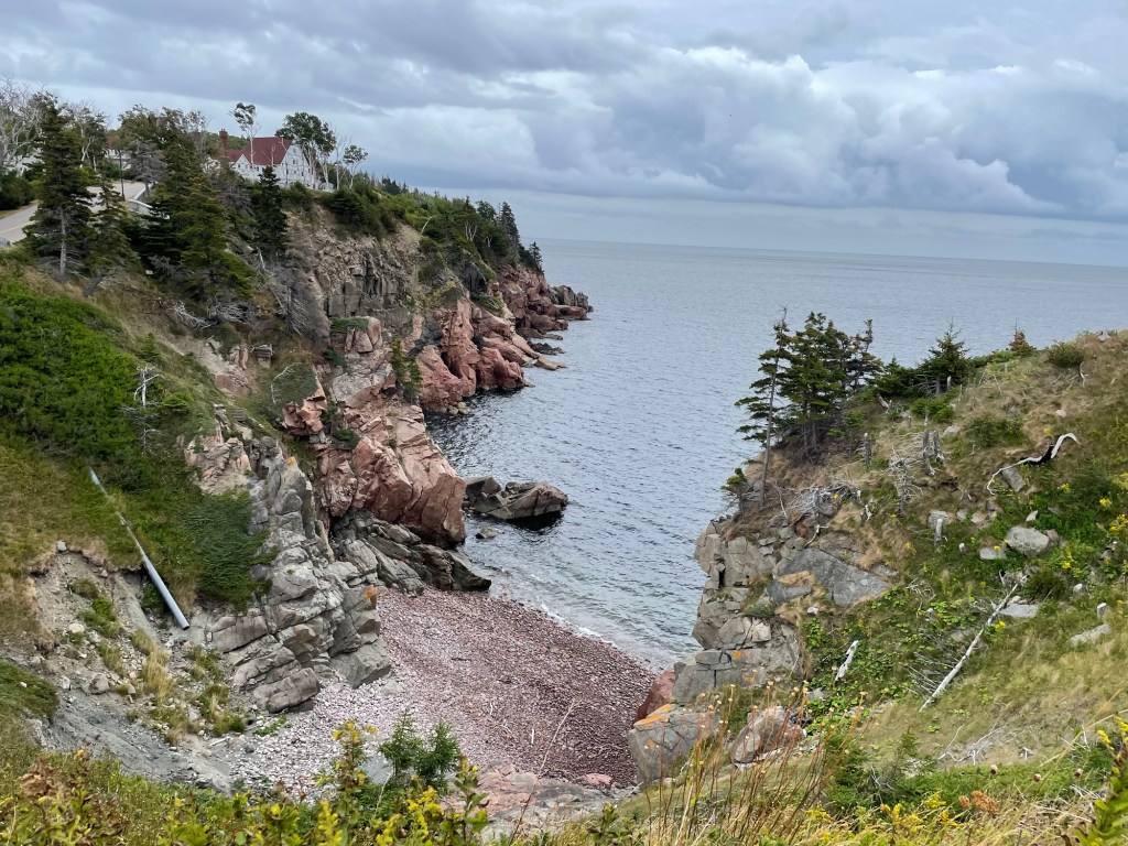  View of a rocky cove from the coastal pedestrian road above, with calm ocean, the cliff wall composed of layers of craggy rocks with some grass and small trees on top, a 2-story house with pitched red roof sitting on top. 