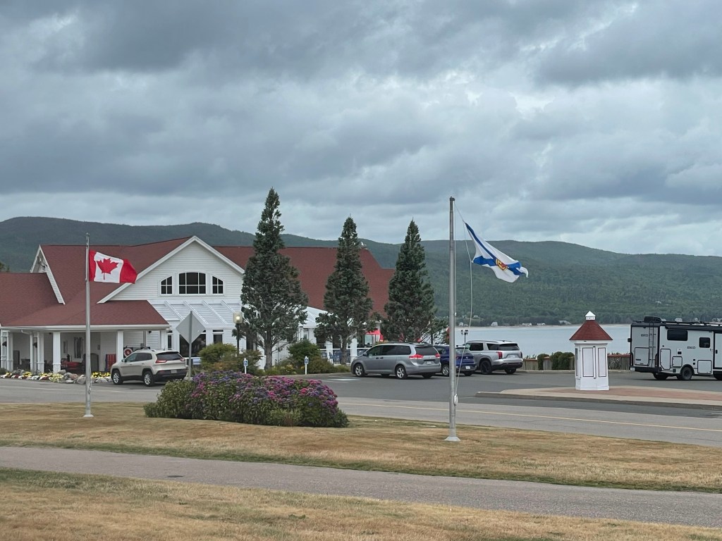 A 2 story white wooden building with red peaked roof stands beside a coastal road, with Canadian and Nova Scotia flags flying on flagpoles in front. Green mountains form the backdrop. 