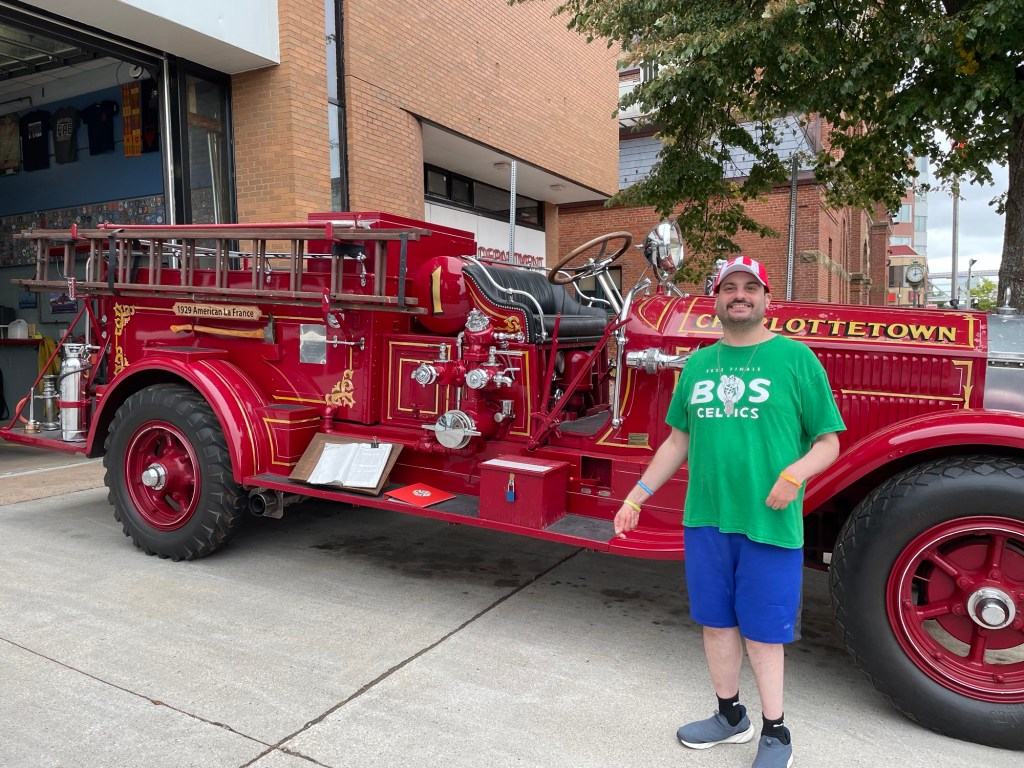 A 30-ish young Caucasian man with a short dark beard stands smiling beside a vintage red fire engine decorated with gold lettering and trim. The truck has large red wheels, polished metal fittings, and a wooden ladder mounted along its side.