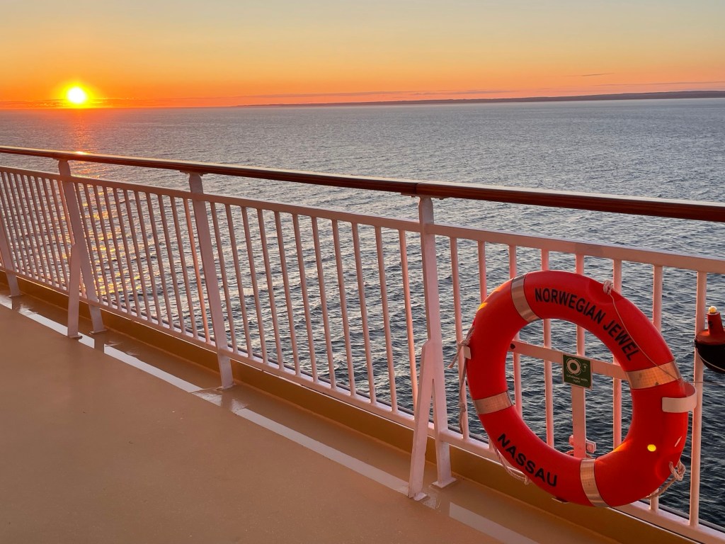 The sun glows just above the horizon, casting a golden-orange reflection across the ocean. A white ship’s railing runs diagonally across the foreground, with a bright red life ring labeled Norwegian Jewel Nassau attached to it.
