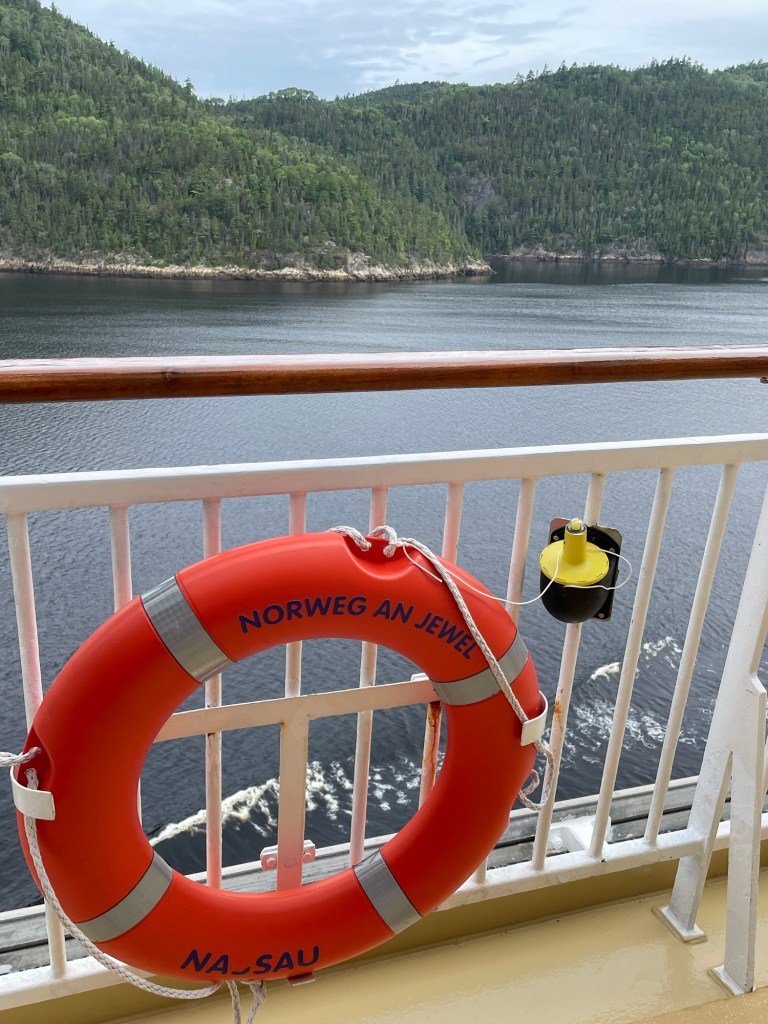 A duo of lush green mountains one in front of the other rise up from a calm flat body of water as viewed from the deck of a cruise ship. Hanging on the rail in the foreground is a large orange life ring labeled with the ships name Norwegian Jewel. 
