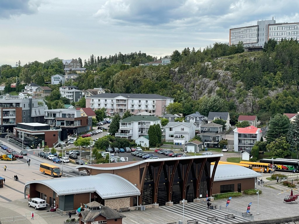A town along one of the mountains along the fjord shows multiple styles of modern mostly 3 story wood or stone buildings, with houses rising on a winding road in the background and a large wide glass building on top of the mountain. 