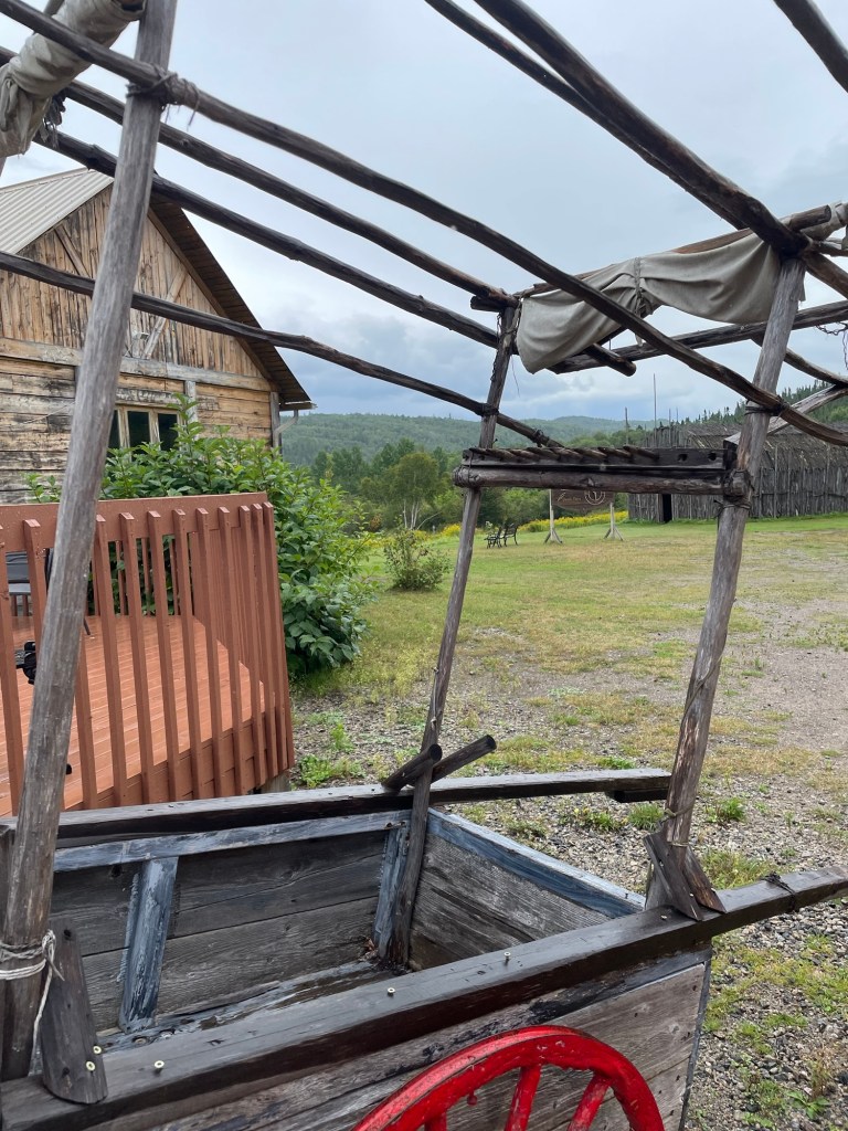 A weathered wooden cart with a red spoked wheel sits beneath a rough wooden frame beside a rustic timber building. Beyond the grassy yard, forested hills rise under a cloudy sky.