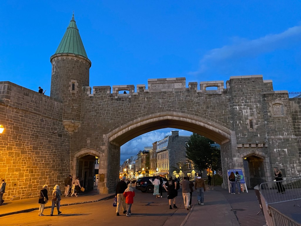 A massive stone gate with a broad central arch and a round tower topped by a green conical roof stands against a vivid blue evening sky. Through the arch, warmly illuminated historic buildings line a busy street where pedestrians stroll. Golden streetlights contrast with the cool twilight beyond the gate.