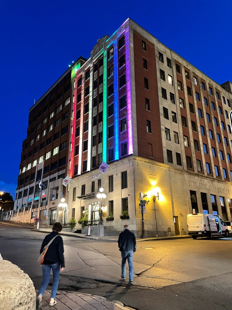 A tall, historic building with red brick and pale stone façades rises against a deep blue evening sky. Vertical strips of red, green, blue, and purple lighting illuminate the central façade. Warm streetlights brighten the entrance as two pedestrians cross the street in the foreground.